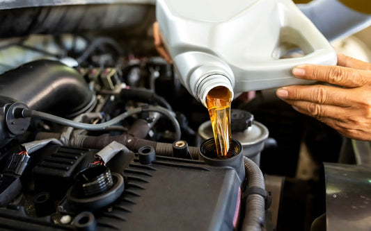 A close-up of a person using both hands to pour amber-colored oil from a silver container into the engine of a vehicle.