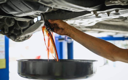 A hand holding an oil plug is pulling away from the undercarriage of a car. Grimy, dark oil is leaking down into a drip pan.