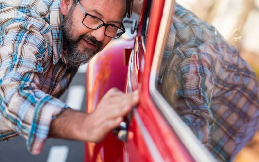 A man with a beard and glasses stands with his face next to the window of a red car. He glides his hand along the paint.