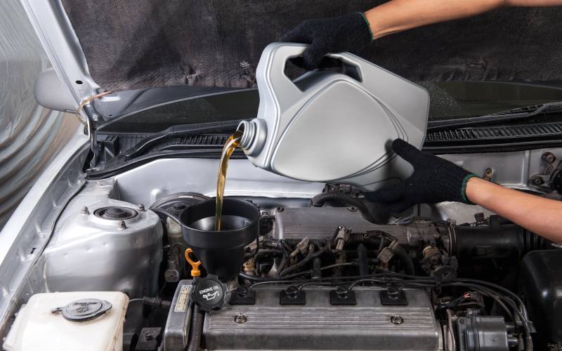 A pair of hands wearing black gloves is dumping a gray container of oil into the open hole of a performance engine.