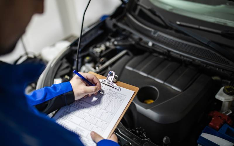 Person standing in front of an engine with an open oil cap. The person has a clipboard and is writing something down.