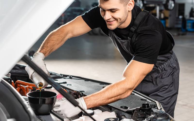 A man in a black shirt and gray overalls is bent over the open hood of a car, pouring oil into a funnel in the engine block.