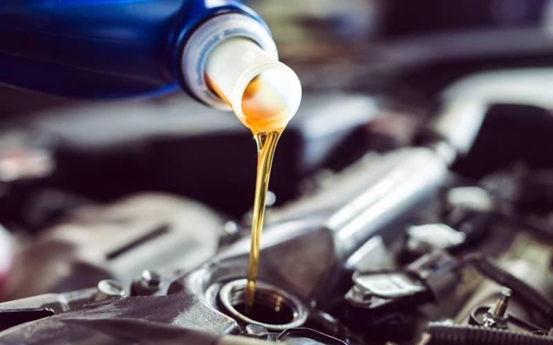 Close-up of the nozzle of a bottle pouring oil into the engine block of a car. The golden oil flows down toward the engine.