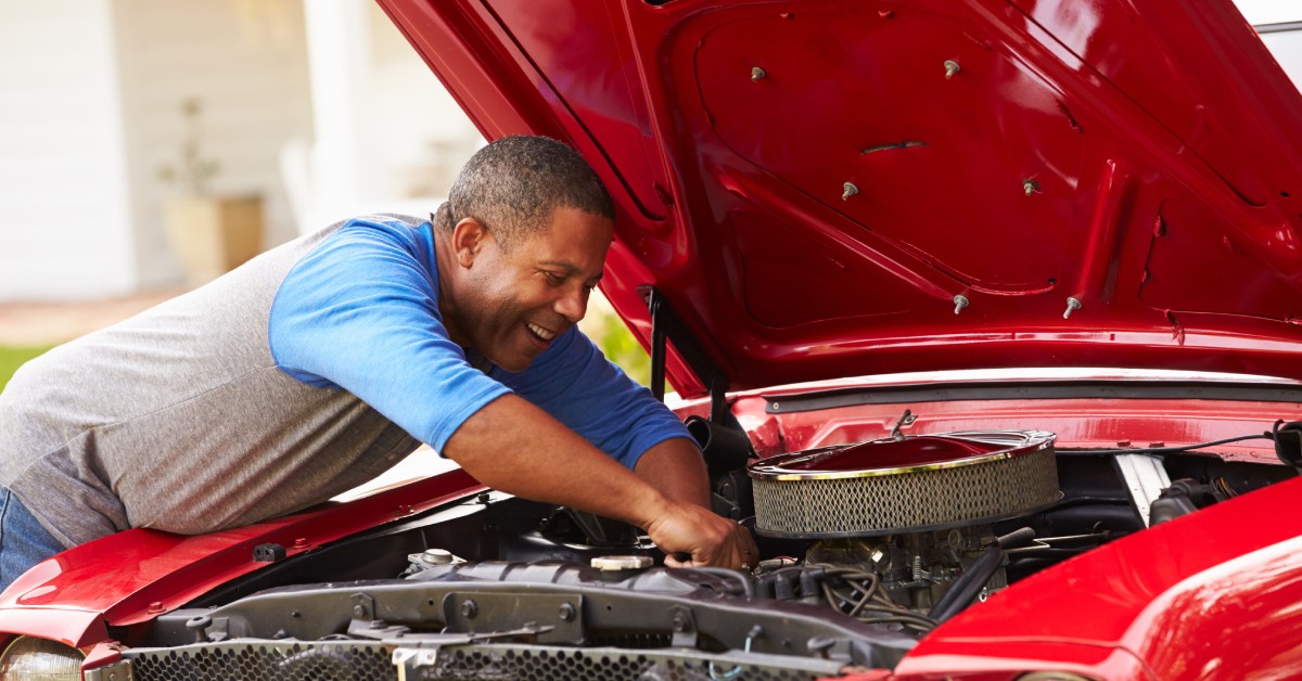 An older man works under the hood of a classic car. The man leans into the engine with a wrench to fine-tune his vehicle.