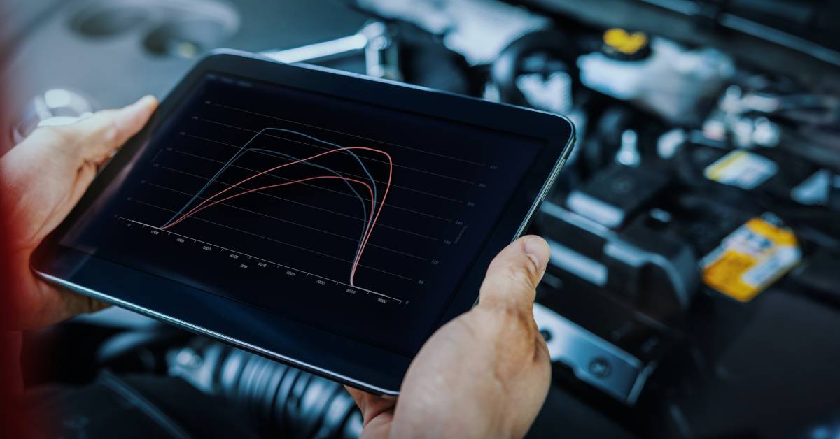 A mechanic in an autobody garage checks an engine's performance using a tablet. The tablet shows lines to assess its progress.