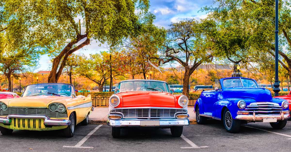 Five classic cars in a parking lot outside. Trees are vibrant green, orange and red, signifying the fall season.