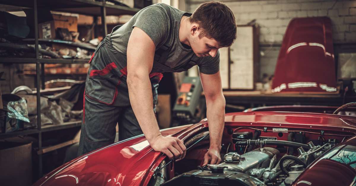 A male mechanic inside an auto shop working on the engine of a red and white classic car. The hood sits on a table nearby.