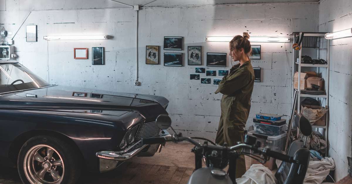 A woman standing in front of a black classic car in a garage surrounded by various vintage memorabilia. She looks pleased.