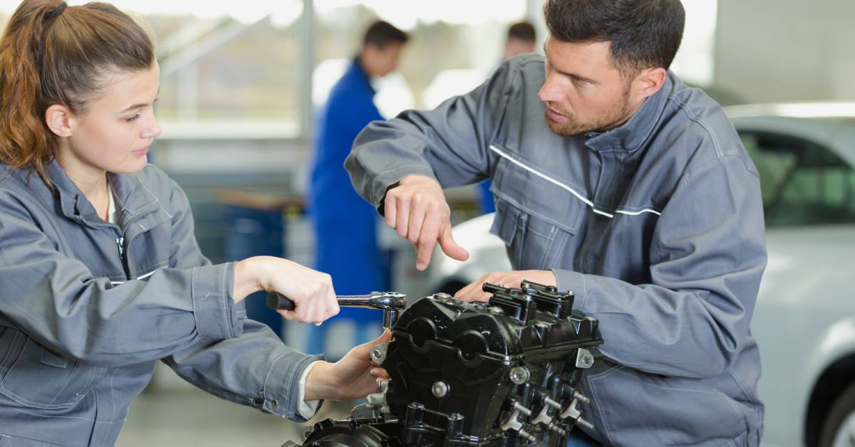 Two mechanics are working together on an engine. One uses a wrench while the other is guiding where to use the tool.