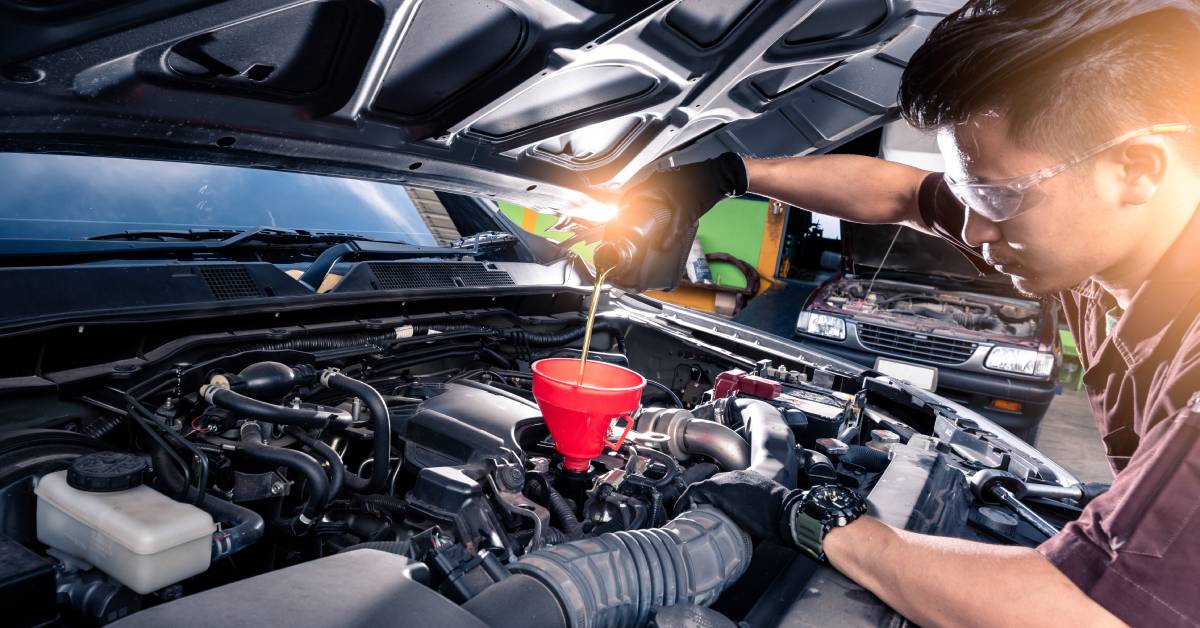 A technician at an autobody shop fills an engine with new lubricant. He wears protective glasses and gloves.