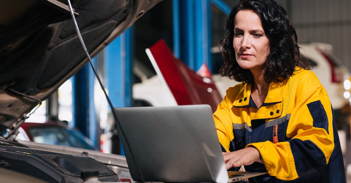 A female mechanic sitting next to a car's open engine. She diagnoses and analyzes the motor's performance with her laptop.