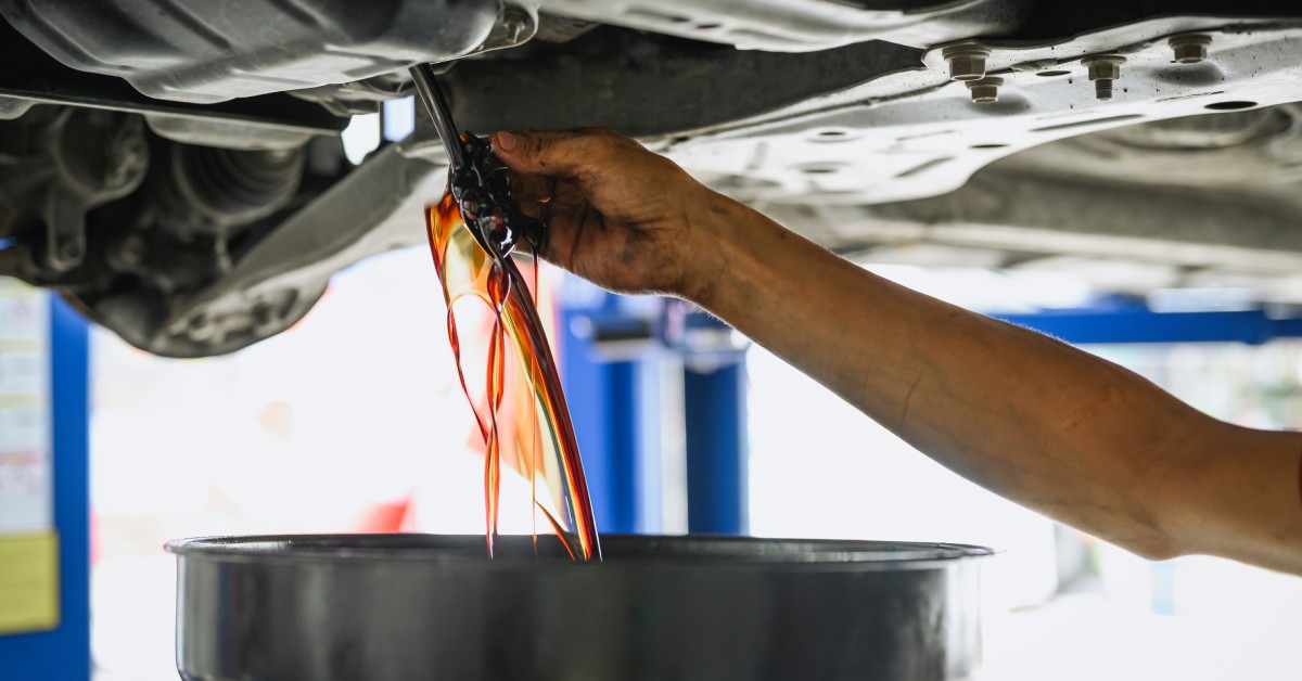 An auto mechanic draining old oil from a vehicle. A cylinder object catches the waterfall of oil coming out of the car.