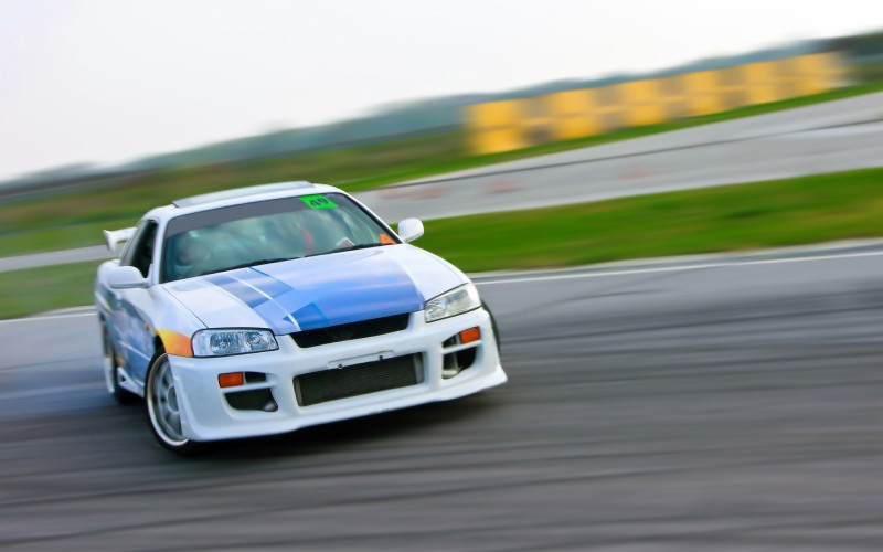 A blurry background shows a white, blue, and orange race car drifting down a race track. The driver turns the car's wheels right on the track.