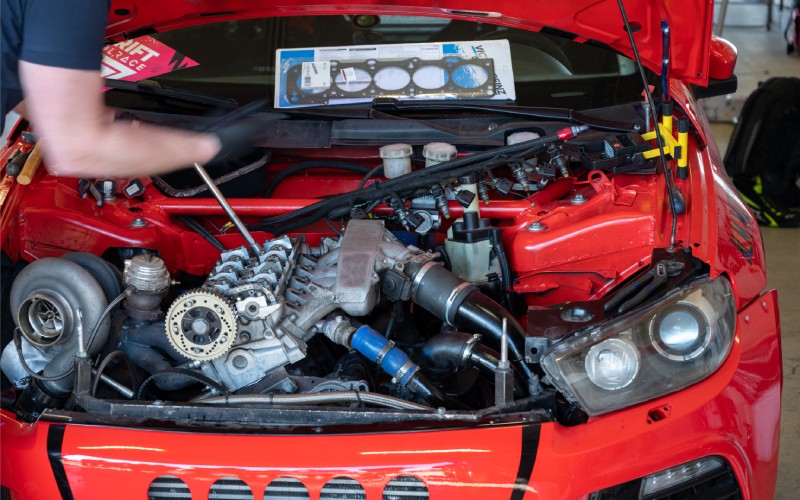 A male car owner works on an upgraded race car engine in a workshop. The man wears gloves to protect his hands.