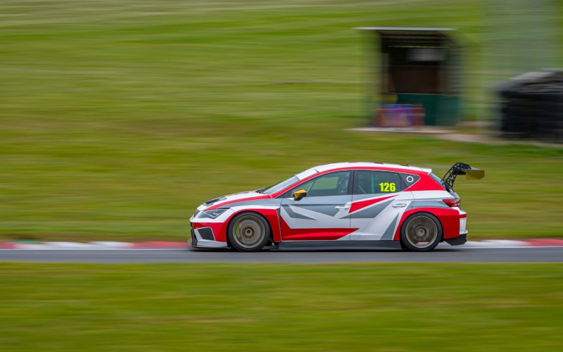 A blurry shot focuses on an orange, gray, and white race car as it circuits a race strip. The car drives straight down the track.