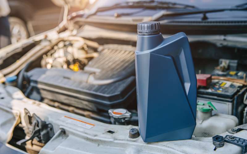 A blue bottle of motor oil sits on the front of a car's hood. The hood is open, revealing the engine parts in the background.