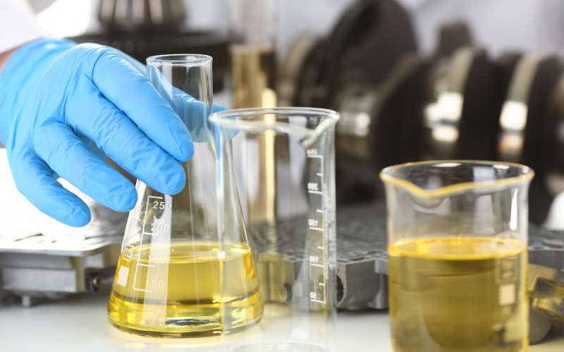 Three test tubes are full of engine oil and face a white background, with a person's hand wearing a protective glove.