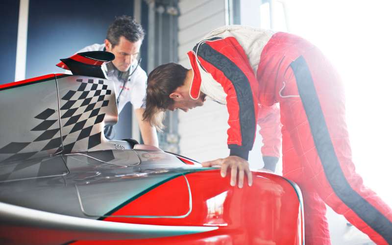 Two race car team members inspect a car engine. One reaches in, and the other peers inside, wearing a headset.