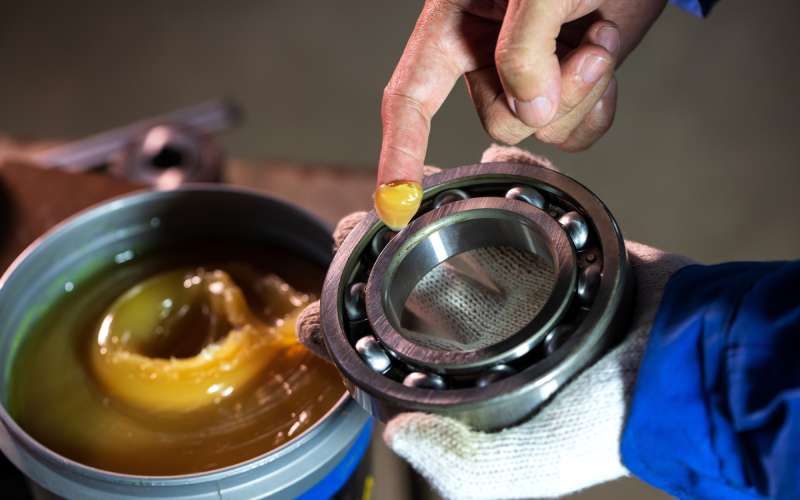 A mechanic with lubricant on their right pointer finger and a ball bearing in their left hand. The background is blurry.