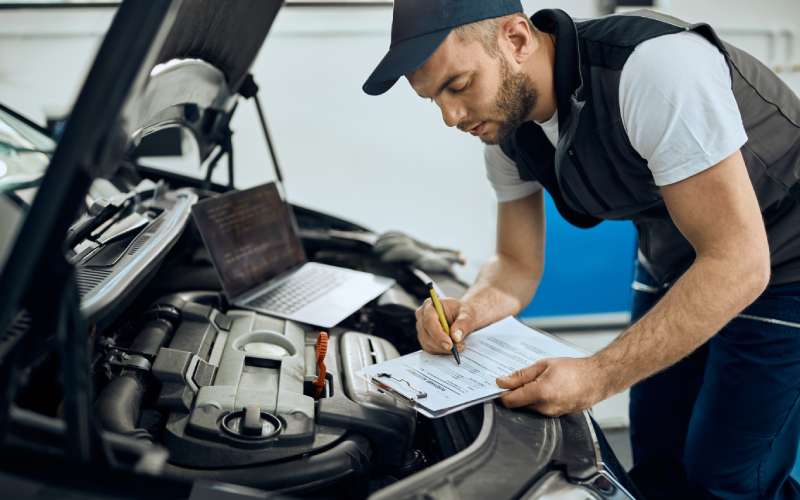 A mechanic standing next to an open car hood holding a clipboard. Inside the hood are the engine and its components.