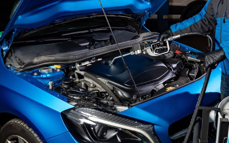 A blue car with its hood open, exposing the engine components inside. A mechanic stands with a pump inside of the engine.