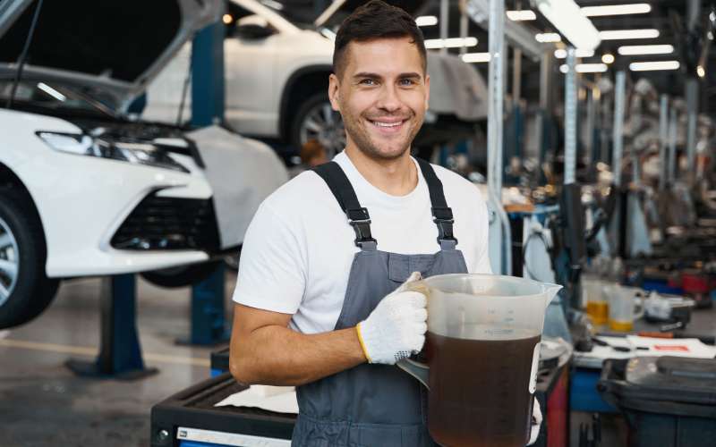 A mechanic wearing overalls stands and smiles while holding a pitcher of engine oil. Raised cars fill the background.