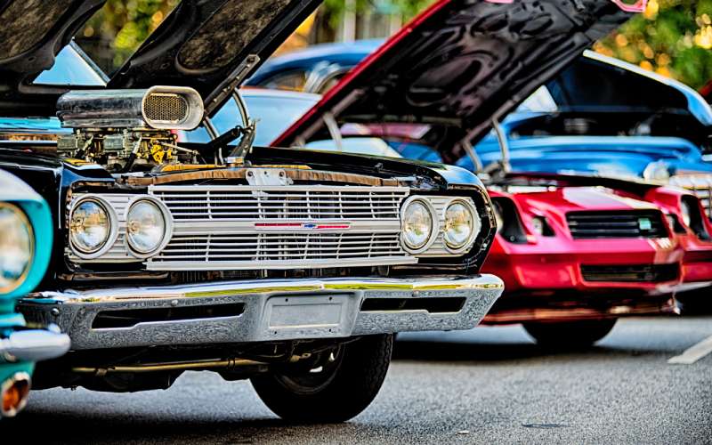 Classic cars parked in a lot at a car show with their hoods up. The second car has a V-type engine.