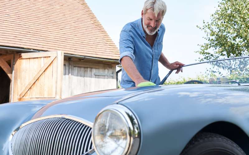 A man in a blue button-down shirt uses a yellow towel to wipe the front of his car. The towel helps restore the car's luster.