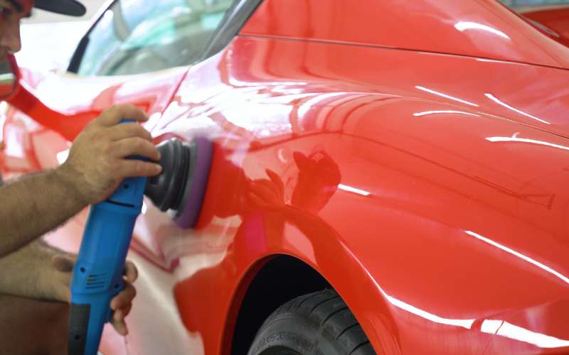 A professional mechanic uses a pad to polish the side of a red classic car. An auto shop is in the car’s reflection.