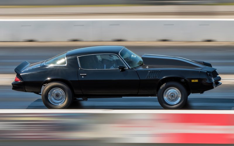 A black drag race car driving down a track fast. A cement wall is in the background of the blurry image.