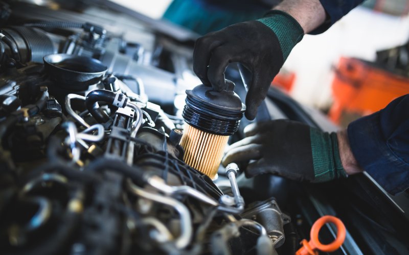 A mechanic removing a dirty oil filter from a car engine. The car's engine fills most of the foreground.