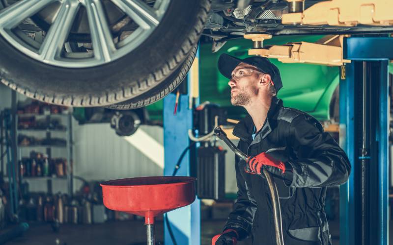 A mechanic holding a tool standing under a car that's lifted up in a garage. There's a large red funnel set up beneath it.