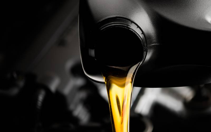 Close-up of the mouth of a black bottle pouring golden oil downward. There's a faintly blurry background with a car.