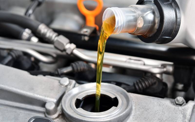 Close-up of a bottle pouring oil into an open hole in a chrome engine. The orange dipstick head is visible in the background.