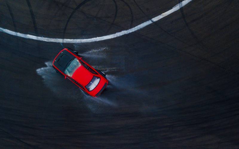 An aerial view of a red race car on a wet, rainy track. The drifting car is kicking up water as it turns.