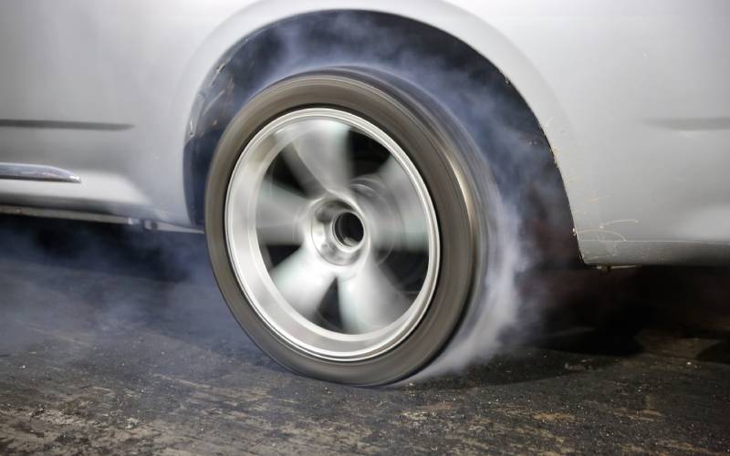 A close-up of a silver car's tire. The wheel is spinning so quickly that it's creating a thick cloud of smoke.