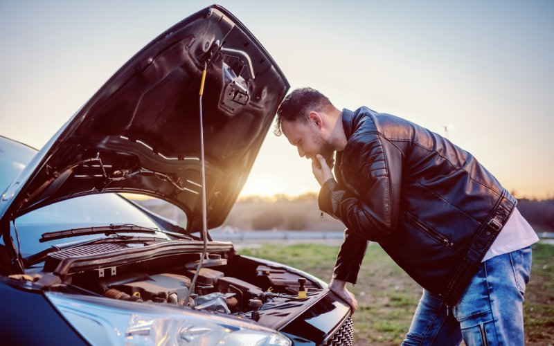 A man in a black leather coat is standing over his car. The hood is up, and he's examining the engine with his hand on his chin.