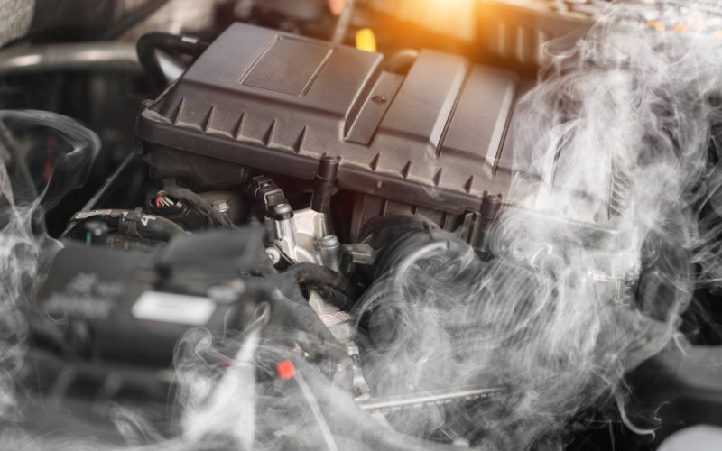  Closeup of a black engine block. An enormous cloud of thick gray smoke is coming from the overheating engine.