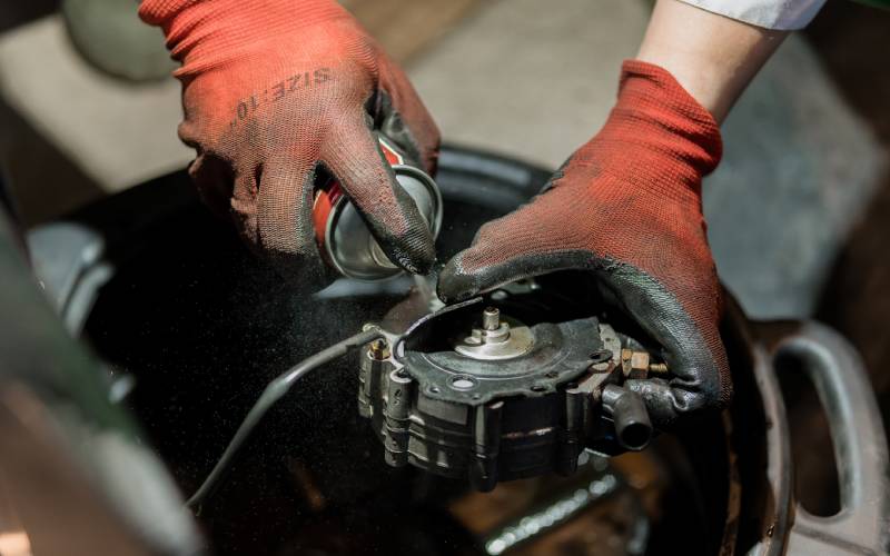 A pair of gloved hands using a spray cannister to apply lubricant to part of an engine. A fine mist comes from the can.