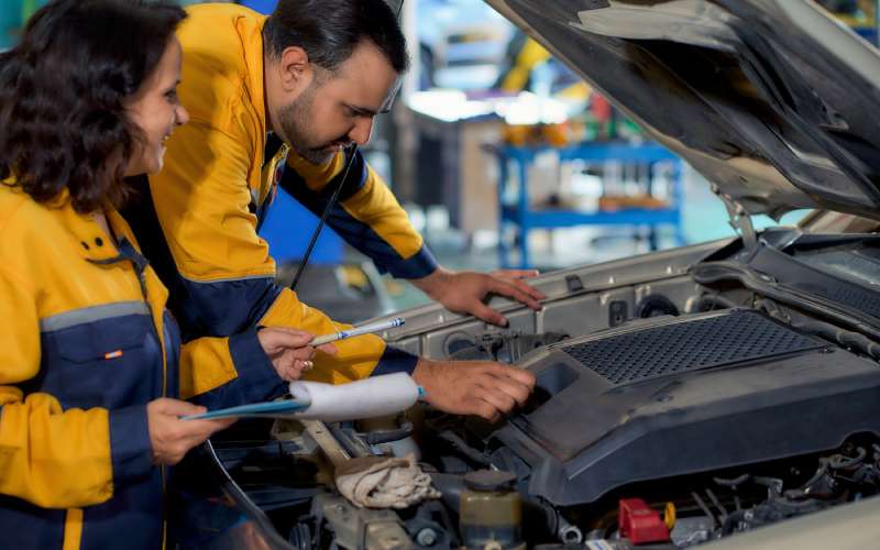 Two mechanics inspect a car's engine. One leans into the engine while the other holds a clipboard and observes.