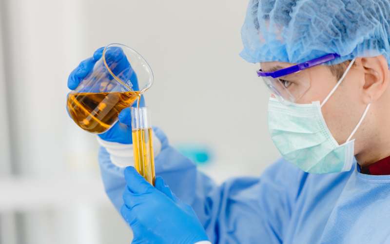 A scientist wearing gloves, a mask, and safety glasses pours synthetic oil from a cylinder into a test tube in a lab.