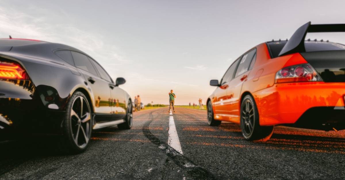 Two drag race car drivers stand at the starting line, awaiting the race starter to signal for them to go. Their drag race cars run effortlessly thanks to drag racing oil.